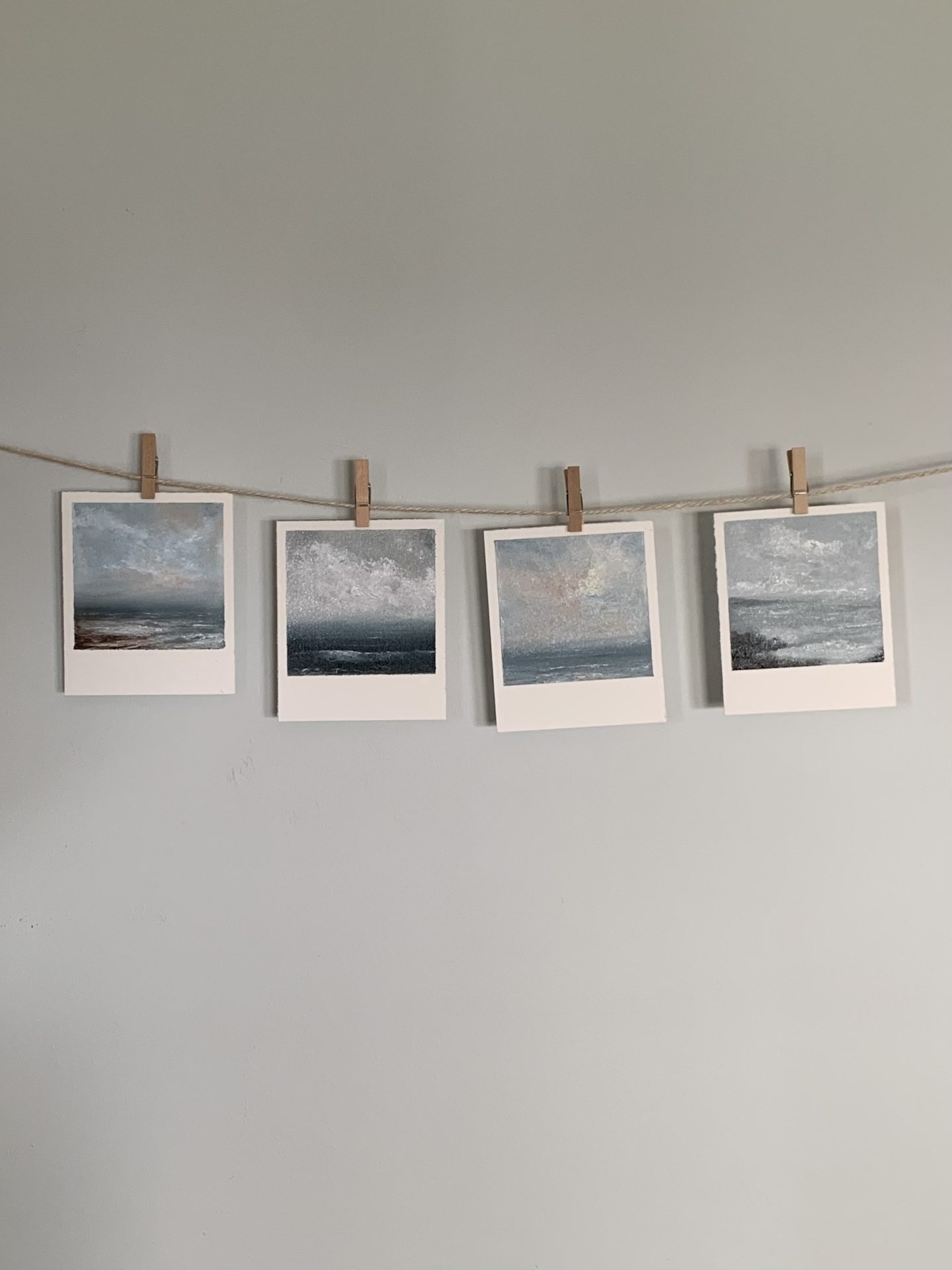 Photo of four original tiny seascape oil paintings by Tisha Mark, Snapshots Series, Nos. 34-37, oil on paper (2023), shown here hanging from tiny clothespins on a line of twine. Paintings are formatted to resemble old-school instant photos.
