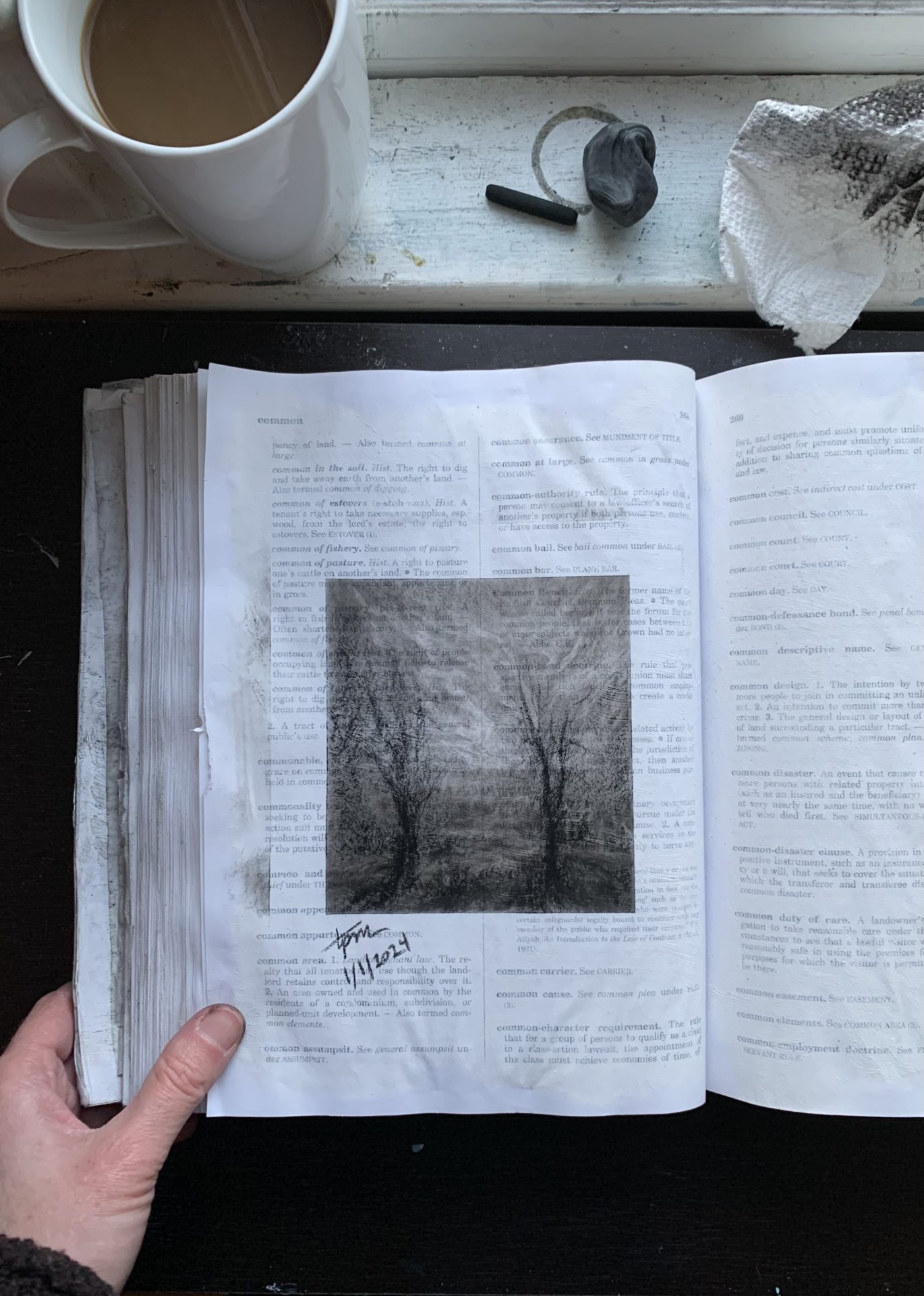 Photo of a law dictionary open to a page with a charcoal landscape sketch in the middle of the page. Printed words on the page are partially visible beneath a thin layer of gesso and the charcoal sketch. A hand holds the page open. On the windowsill at the top of the photo is a cup of coffee, a piece of charcoal, an eraser, and a paper towel smudged with charcoal.