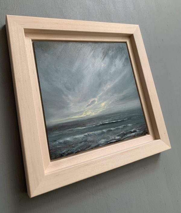 Original seascape oil painting by Tisha Mark, "The Watching" 6"x6" oil on gessobord (2024). Light emerges in a stormy sky over a stormy sea. Shown here in an unfinished maple frame at an angle from left.