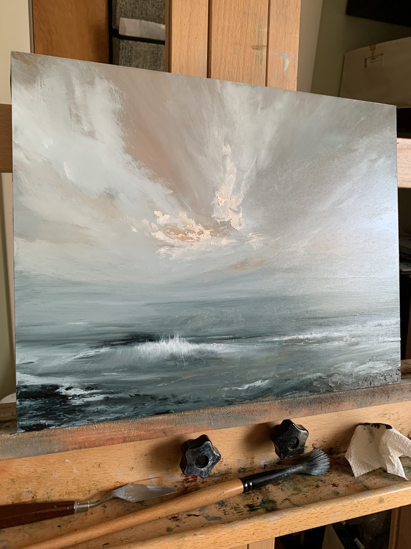 Photo of an original oil painting by Tisha Mark, "A Little Light, At Last" 11"x14" oil on cradled gessobord (2024). Light emerges in an orange toned stormy sky over a stormy sea. Shown here on an easel.