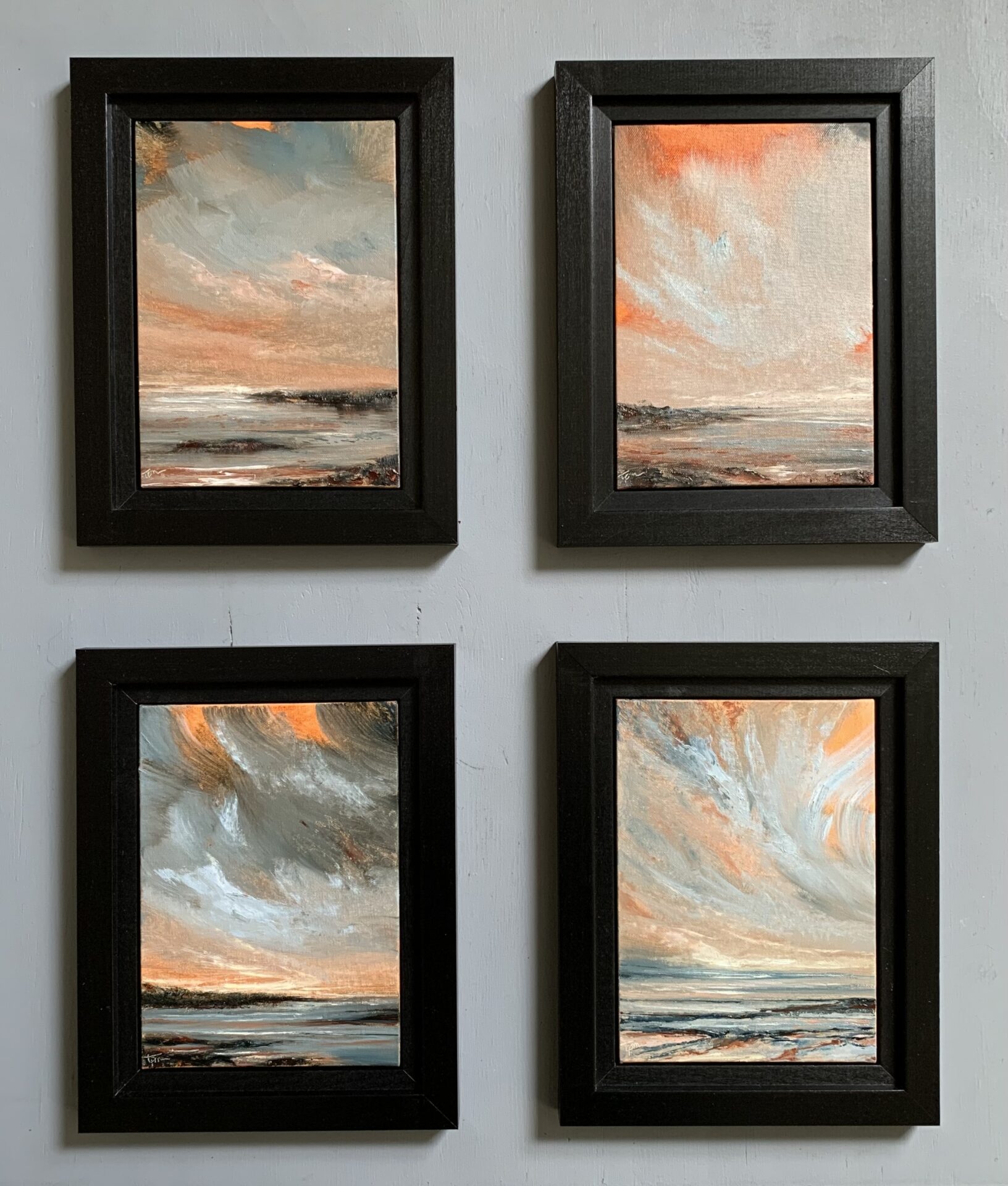 Photo of four original seascape oil paintings by Tisha Mark. The paintings feature an orange-toned sunset sky with cloud formations, and light reflecting in the water of a rocky coast below. Shown here in black frames against a gray background.
